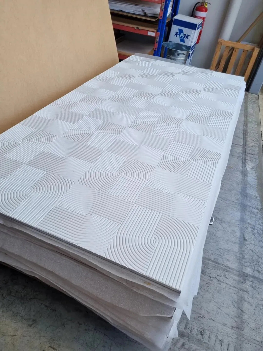 Stack of white decorative plywood panels with geometric textured patterns in a warehouse.
