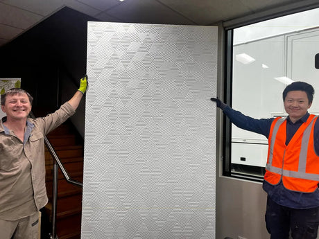 Two workers holding a decorative patterned plywood panel indoors next to a window