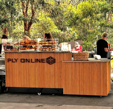 Outdoor plywood food stall with stacked pastries, forest backdrop, and people serving customers