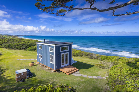 Modern blue tiny house with plywood cladding by the ocean, surrounded by lush greenery and clear sky