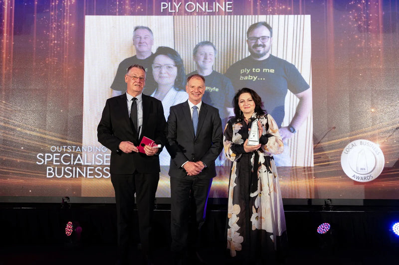 Three people stand on a stage in front of a large screen at an awards event. The screen shows a group photo and the text 'PLY ONLINE' and 'OUTSTANDING SPECIALISED BUSINESS.