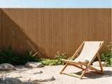 A light natural wood folding deck chair with a beige canvas sling sits on a gravel patio in front of a tall bamboo slat fence with green plants.